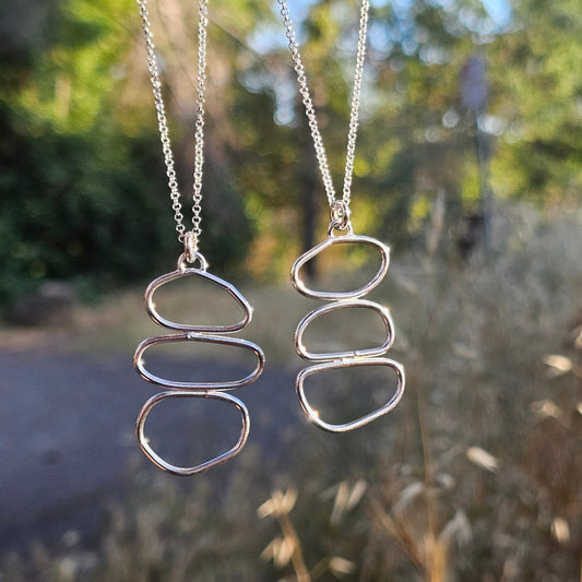 Two silver necklaces with pebble-like pendants hanging against a natural background.